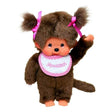 Officially licensed Monchhichi girl plush doll with dark brown fur and hair styled into two pigtails with bright pink ribbons. The doll has a freckled vinyl face, a long tail, and is wearing a white bib with a pink checkered border that reads "Monchhichi".
