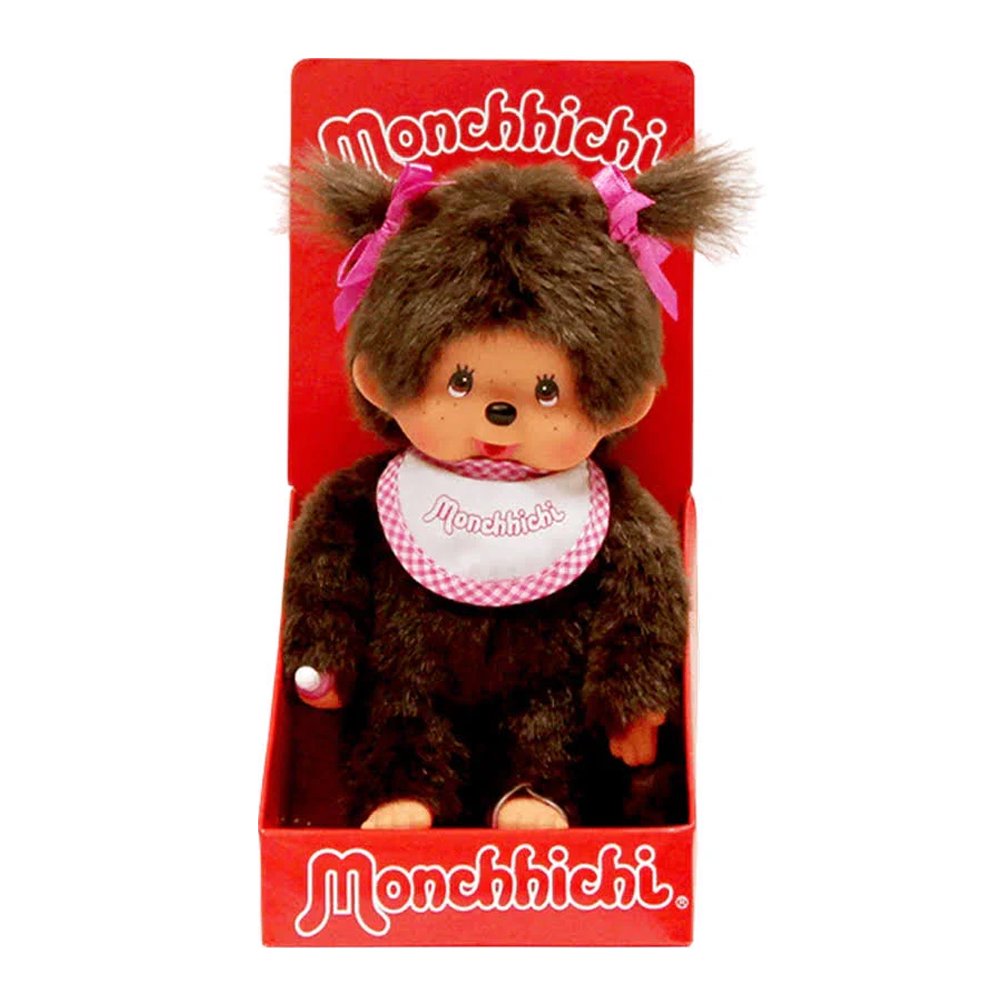 Officially licensed Monchhichi girl plush doll with dark brown fur and hair styled into two pigtails with bright pink ribbons. The doll has a freckled vinyl face, a long tail, and is wearing a white bib with a pink checkered border that reads "Monchhichi".
