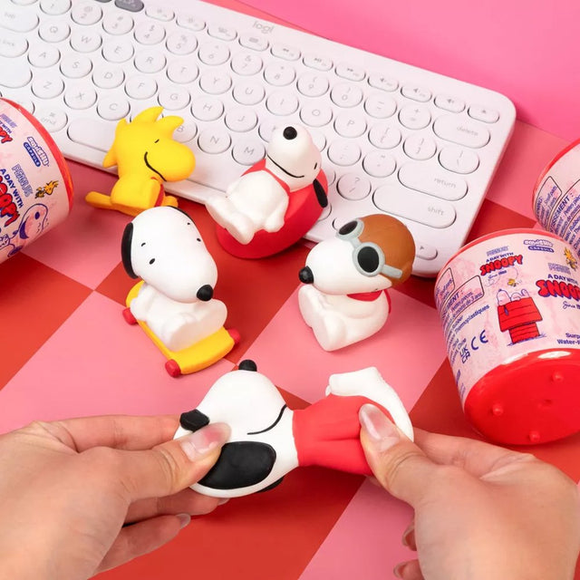 Officially licensed Peanuts Snoopy and Woodstock Squishu water-filled squishy figures in various poses displayed alongside a red surprise capsule