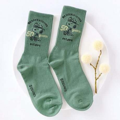 A set of five crew-length socks in black, beige, white, green, and mustard yellow. Each sock features an embroidered graphic of Snoopy as a Beagle Scout leader.