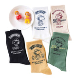 A set of five crew-length socks in black, beige, white, green, and mustard yellow. Each sock features an embroidered graphic of Snoopy as a Beagle Scout leader.