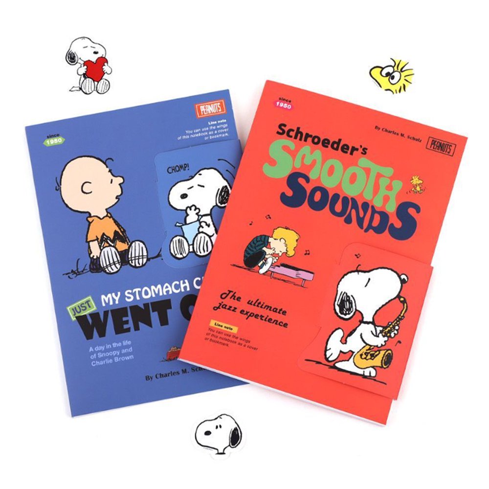 Two officially licensed Peanuts notebooks in red and blue featuring Snoopy and Charlie Brown. The image demonstrates the unique "wing" cover flaps being used as bookmarks and page markers within the lined journal pages.