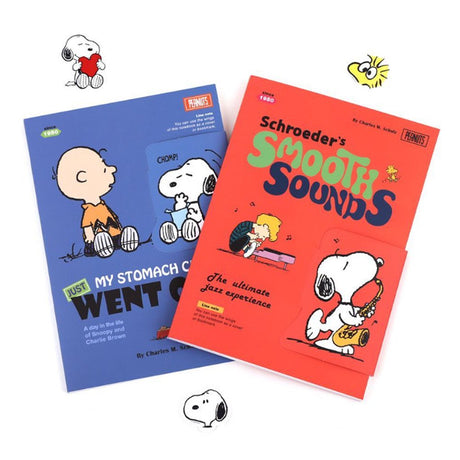 Two officially licensed Peanuts notebooks in red and blue featuring Snoopy and Charlie Brown. The image demonstrates the unique "wing" cover flaps being used as bookmarks and page markers within the lined journal pages.