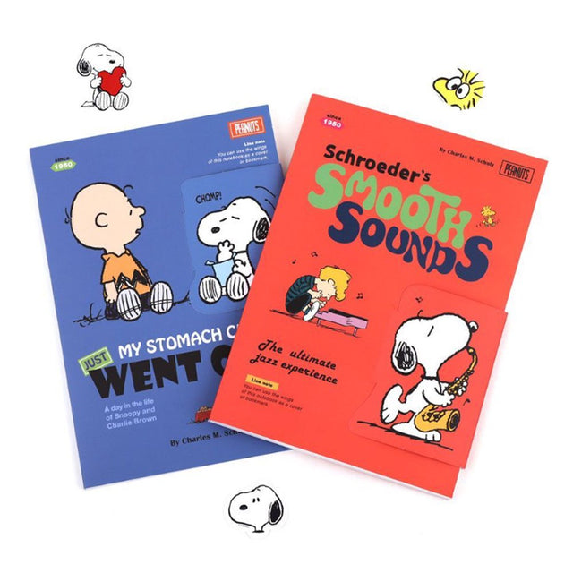 Two officially licensed Peanuts notebooks in red and blue featuring Snoopy and Charlie Brown. The image demonstrates the unique "wing" cover flaps being used as bookmarks and page markers within the lined journal pages.