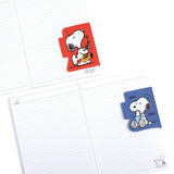 Two officially licensed Peanuts notebooks in red and blue featuring Snoopy and Charlie Brown. The image demonstrates the unique "wing" cover flaps being used as bookmarks and page markers within the lined journal pages.