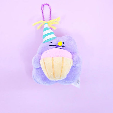 A Pokémon Ditto plush keychain holding a celebration cupcake and wearing a striped party hat with yellow yarn on top, attached to a colorful ball chain