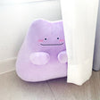 An officially licensed Pokémon Ditto plush toy in a vibrant light purple color. The plush features a simple smile, and three pink diagonal lines on each cheek to represent blushing.