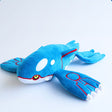 A large, horizontal plush of the Legendary Pokémon Kyogre. The plush is vibrant blue with white oval patches on its head and large, flat flippers featuring red circular patterns and white finger-like tips. It has small yellow eyes and a sleek, aerodynamic body shape.