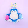 A Pokémon Snorlax plush keychain holding a celebration cupcake and wearing a striped party hat with yellow yarn on top, attached to a colorful ball chain