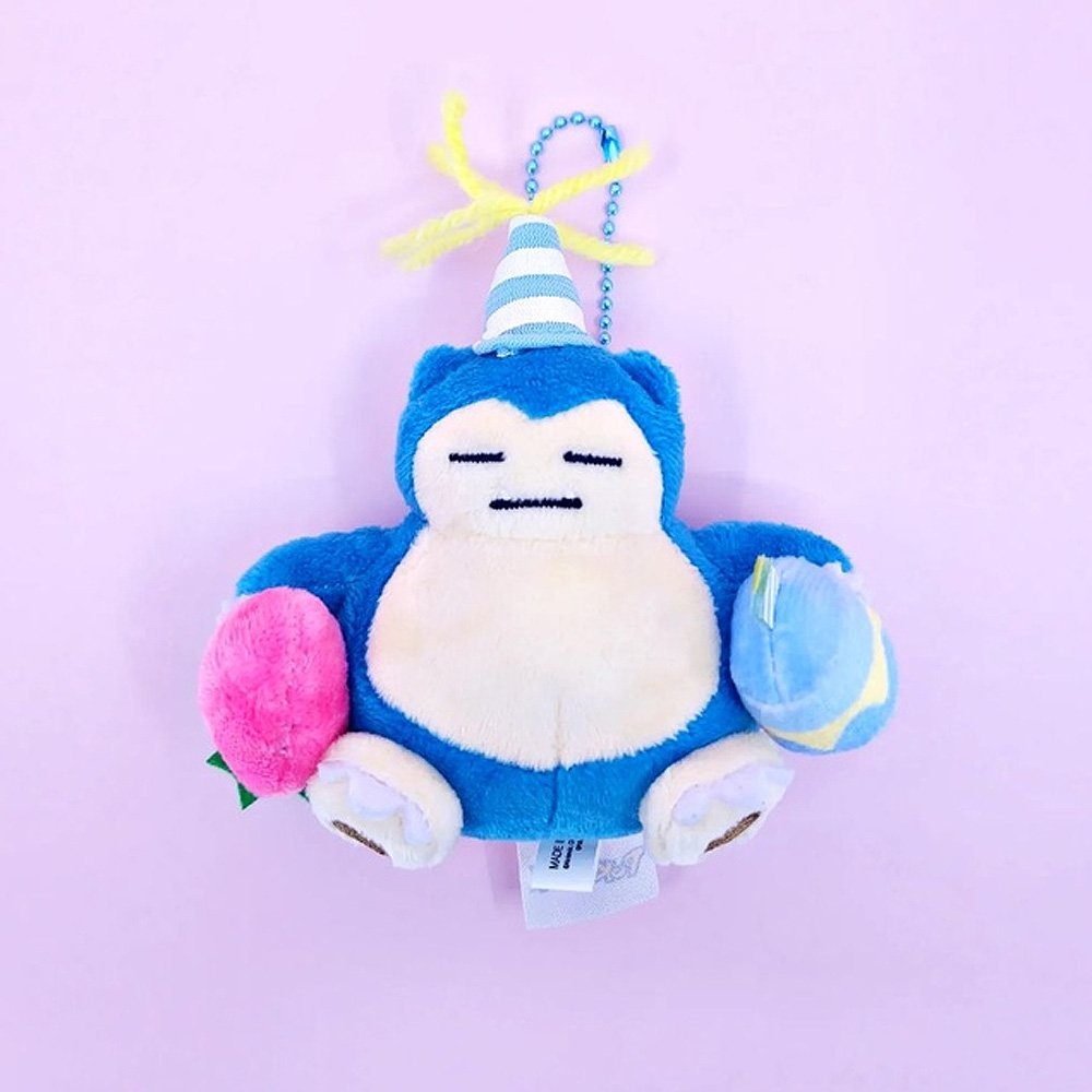 A Pokémon Snorlax plush keychain holding a celebration cupcake and wearing a striped party hat with yellow yarn on top, attached to a colorful ball chain