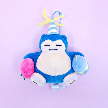 A Pokémon Snorlax plush keychain holding a celebration cupcake and wearing a striped party hat with yellow yarn on top, attached to a colorful ball chain