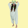 Official Pokémon Sprigatito Kids Costume Kigurumi Onesie Jumpsuit worn by a smiling young model holding the hooded ears. Features a soft pale mint-green fleece body, green leaf collar, and a Sprigatito face hood with pink eyes. Ideal for kids' Halloween costumes, cosplay party suits, sleepovers, and holiday gifts