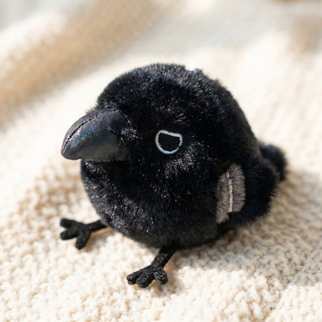 A round, fluffy black crow plush keychain features a large shiny dark blue beak, small embroidered eyes, tiny grey wing patches, and small black feet sticking out from its spherical body.