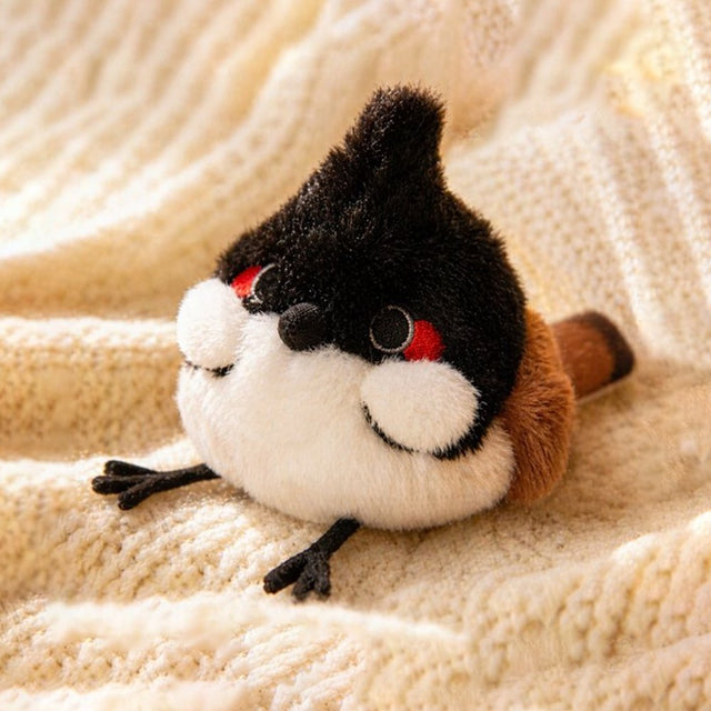 An adorable, round red-whiskered bulbul bird plush keychain with a pointed black crest and a plump body. The plush features striking red and white cheek patches, small embroidered eyes, brown wings, and tiny black feet extended forward.