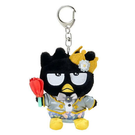A plush keychain of the Sanrio character Badtz-Maru, a black penguin with a grumpy expression. He is wearing a golden crown with a white flower, a silver tuxedo jacket with gold accents, and floral pants. He holds a red rose in one hand and is attached to a silver metal lobster clasp.