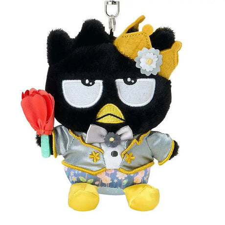 A plush keychain of the Sanrio character Badtz-Maru, a black penguin with a grumpy expression. He is wearing a golden crown with a white flower, a silver tuxedo jacket with gold accents, and floral pants. He holds a red rose in one hand and is attached to a silver metal lobster clasp.