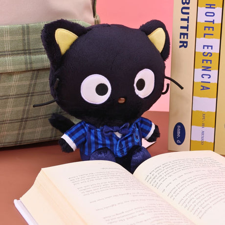 Sanrio officially licensed Chococat plush doll seated on a desk in front of an open book and stacked textbooks. Chococat is dressed in a school uniform featuring a blue striped shirt, a white collar, and a bow tie. The collectible plush is defined by its black fur, yellow inner ears, and large white eyes.