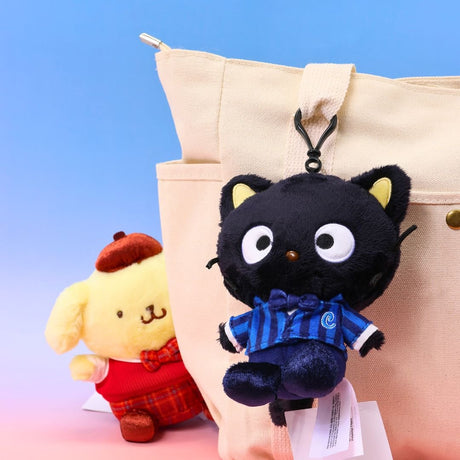 Sanrio official plush keychains from the school uniform series. Chococat is hanging from a beige canvas tote bag handle, wearing a blue striped suit. Pompompurin is seated on the left, wearing a red sweater vest and a brown beret. Ideal as collectible bag charms and accessories.
