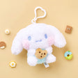 A high-texture, shaggy white Cinnamoroll plush keychain holding a small brown chocolate chip cookie. The character has blue embroidered eyes, pink blushed cheeks, and a mint-green outfit, attached to a white heart-shaped plastic clip.