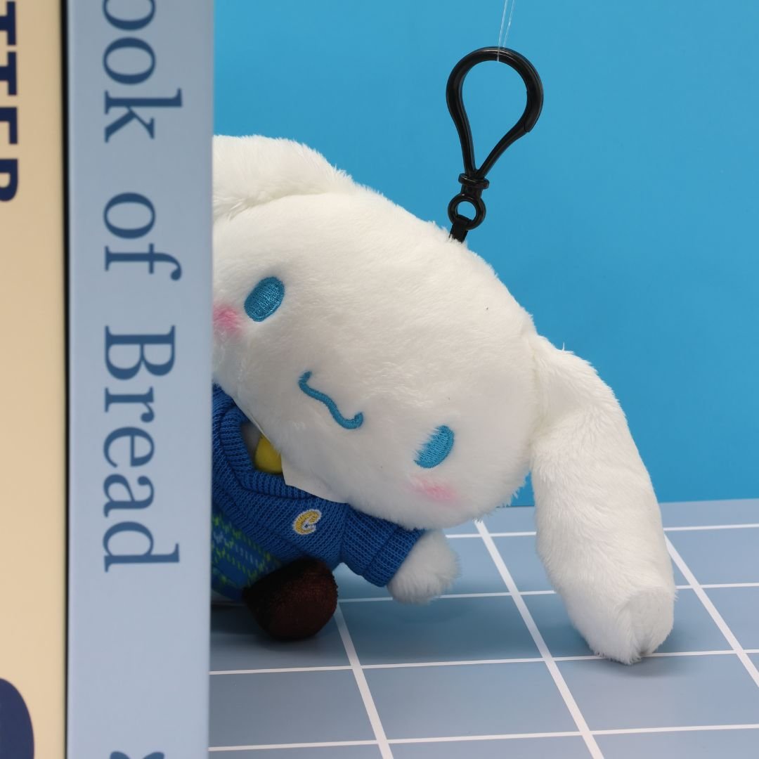 Sanrio officially licensed Cinnamoroll Clip On plush doll dressed in a school uniform. He wears a blue plaid skirt, a V-neck blue sweater, a yellow tie, and behind a books with colorful spines. The soft, white plush features Cinnamoroll's iconic blue-embroidered eyes and swirling mouth, making it a perfect collectible toy.