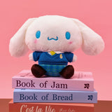 Sanrio officially licensed Cinnamoroll plush doll dressed in a school uniform. He wears a blue plaid skirt, a V-neck blue sweater, a yellow tie, and is seated on a stack of books with colorful spines. The soft, white plush features Cinnamoroll's iconic blue-embroidered eyes and swirling mouth, making it a perfect collectible toy.