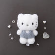 A fluffy white Dear Daniel plush doll keychain wearing textured grey overalls with a heart patch, centered on a dark grey background surrounded by small decorative plastic hearts and stars.
