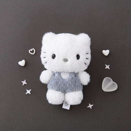 A fluffy white Dear Daniel plush doll keychain wearing textured grey overalls with a heart patch, centered on a dark grey background surrounded by small decorative plastic hearts and stars.
