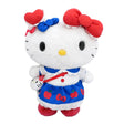 An officially licensed Hello Kitty plush wearing a blue apple-print skirt with a white lace hem, a white shirt with a blue collar, and a red crossbody bag. She has multiple red and blue bows on her head and is wearing red shoes.