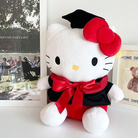 An officially licensed Sanrio Hello Kitty plush toy dressed in a black graduation cap with a gold tassel and a matching gown featuring a large red bow