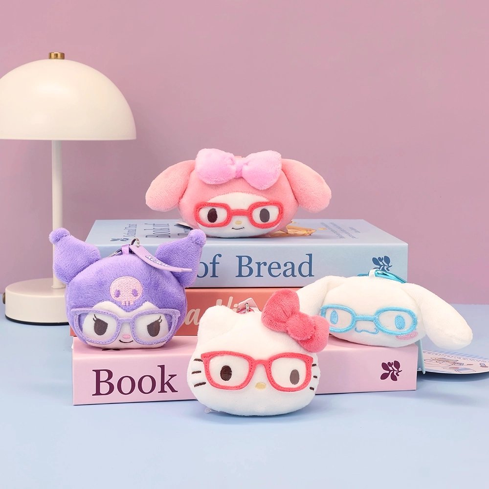 Sanrio plush keychains of Hello Kitty, My Melody, Kuromi, and Cinnamoroll mascot heads, each wearing glasses, are arranged on a stack of books next to a white desk lamp. This styled photo showcases the collectible bag charms.