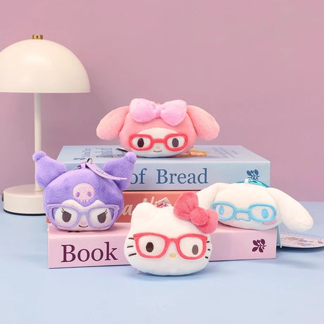 Sanrio plush keychains of Hello Kitty, My Melody, Kuromi, and Cinnamoroll mascot heads, each wearing glasses, are arranged on a stack of books next to a white desk lamp. This styled photo showcases the collectible bag charms.