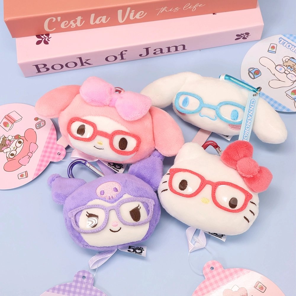 Sanrio plush keychains of Hello Kitty, My Melody, Kuromi, and Cinnamoroll mascot heads, each wearing glasses, are arranged on a stack of books next to a white desk lamp. This styled photo showcases the collectible bag charms.