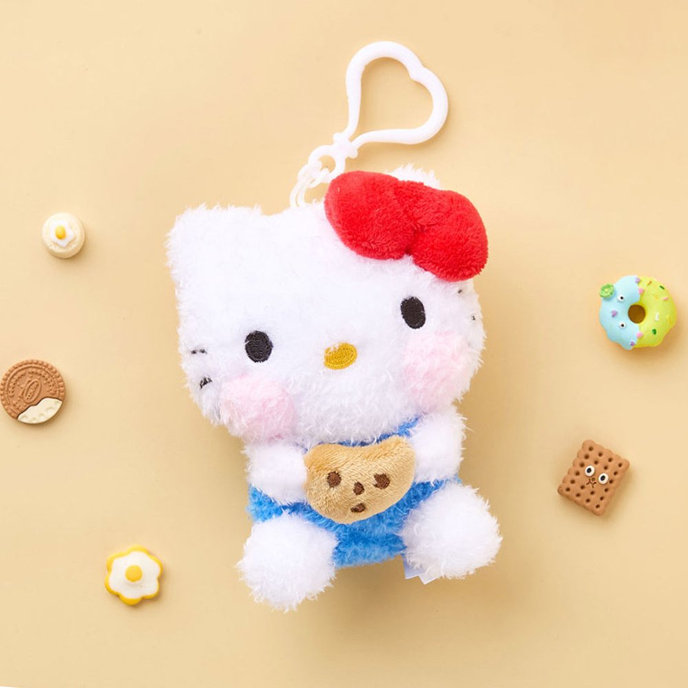 A high-texture, shaggy Hello Kitty plush keychain holding a small brown chocolate chip cookie. She is wearing a red bow and blue overalls with pink blushed cheeks, attached to a white heart-shaped plastic clip.