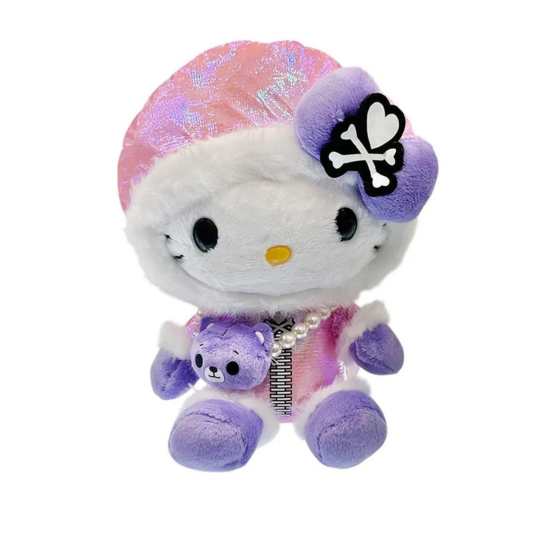  Hello Kitty x Tokidoki design is perfect for the holiday season. Hello Kitty sports a trendy look with an iridescent pink holographic beanie adorned with her signature purple bow decorated with a white heart and crossbones skull Tokidoki mark. She's dressed in a stylish pink holographic vinyl puffer jacket and holds an adorable purple bear plush with a pearl chain strap, giving her a fashionable streetwear vibe. Her soft plush body in white and purple creates the perfect bean doll silhouette for displaying