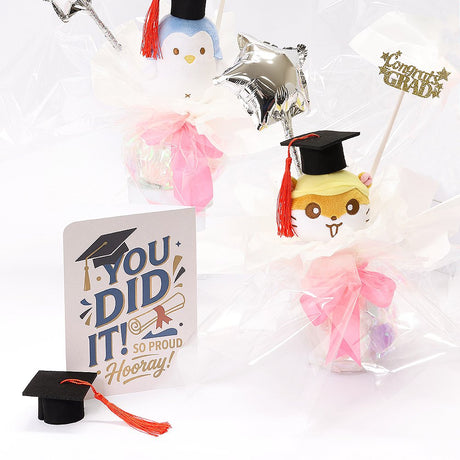 An officially licensed Class of 2026 Sanrio graduation gift set featuring six popular plush characters, Usahana, Corocorokuririn, Wish Me Mell, and Dinguri Days Mashu wearing black graduation caps. The characters are arranged like a bouquet with silver star balloons and "Congrats Grad" decorations.