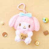 A high-texture, shaggy pink My Melody plush keychain holding a small brown chocolate chip cookie. She features a blue bow, rosy pink cheeks, and is attached to a pink heart-shaped plastic clip.