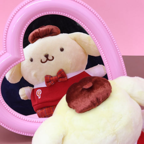 Pompompurin Sanrio plush doll in a formal red school uniform, including a sweater vest, plaid bow tie, and matching hat (beret), admiring his reflection in a pink heart-shaped mirror. The plush is a highly collectible Sanrio school uniform series toy.