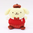 Officially licensed Sanrio Pompompurin wears a red knitted sweater vest, a plaid bow tie, matching plaid shorts, and a brown beret hat. 