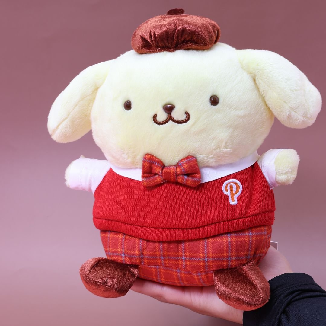 Officially licensed Sanrio Pompompurin plush doll, held in hands, Pompompurin wears a red knitted sweater vest, a plaid bow tie, matching plaid shorts, and a brown beret hat. 