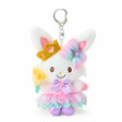 A plush keychain of the Sanrio character Wish me mell, a white bunny with pink-and-white polka dot patterns inside her ears. She is wearing a golden crown, a large rainbow-gradient bow, and a lavender-and-pink princess dress with a floral organza skirt. She holds a yellow flower and is attached to a silver metal lobster clasp.
