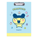 Two officially licensed Tamagotchi clipboards: one light blue featuring Mametchi with cupcakes and sweets, and one pale yellow featuring Kuchipatchi with stars and snacks. Both clipboards include a silver metal clip and the official Tamagotchi logo.