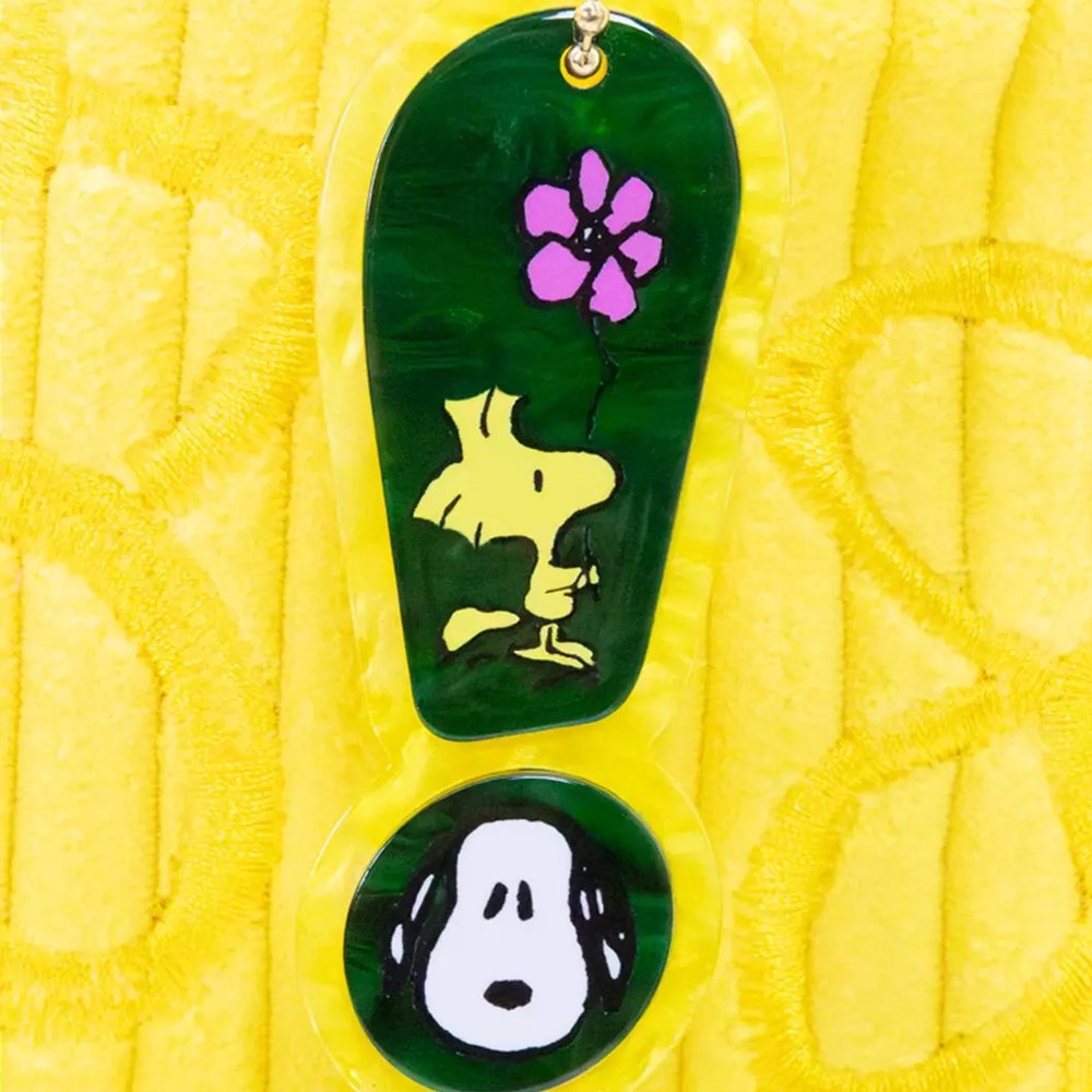 A bright yellow corduroy tote bag shaped like Snoopy's head with long, floppy green ears, subtle embroidered yellow flowers on the face, and a hanging black Peanuts keychain charm

