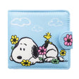 A light blue bi-fold wallet featuring Snoopy and Woodstock relaxing among colorful flowers, shown next to its open view displaying a bright yellow interior with card slots and a clear ID window.