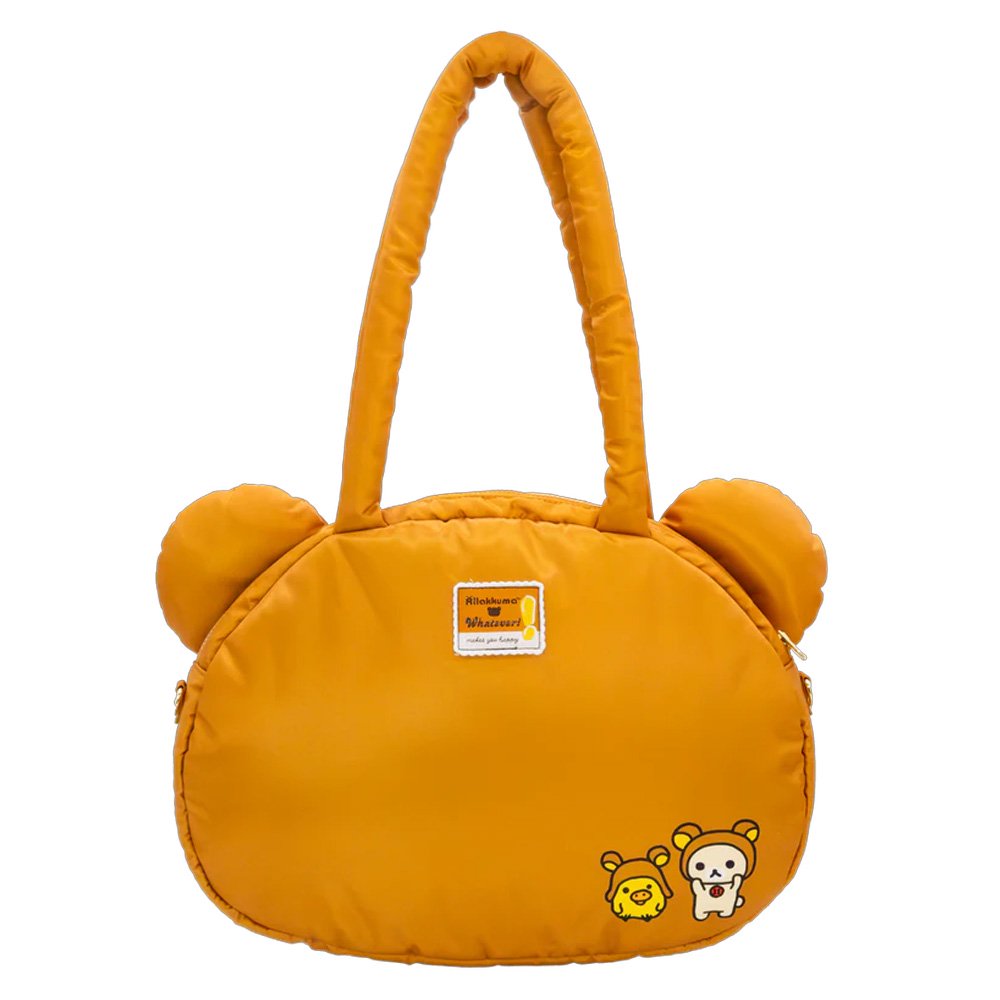 An officially licensed Rilakkuma puffy face crossbody bag in a brown color. It has a white muzzle with a brown nose and mouth, large dark eyes, and long padded shoulder straps. A small yellow exclamation point charm hangs from the base of one handle.