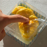 Finger pressing down on a soft golden croissant squishy inside a clear plastic bakery gift box, demonstrating soft squishy foam resistance for stress relief and anxiety relief
