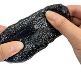 Both hands pulling and stretching a dark charcoal Stone Crunch Squishy Slime Toy revealing a bumpy rock-like surface and dense ASMR crunch beads for tactile anxiety relief