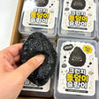 Hand holding and squeezing a black Charcoal Stone Crunch Squishy Slime Toy over clear plastic storage cases with cute Korean stone character labels, ideal stress relief sensory fidget toy and holiday stocking stuffer