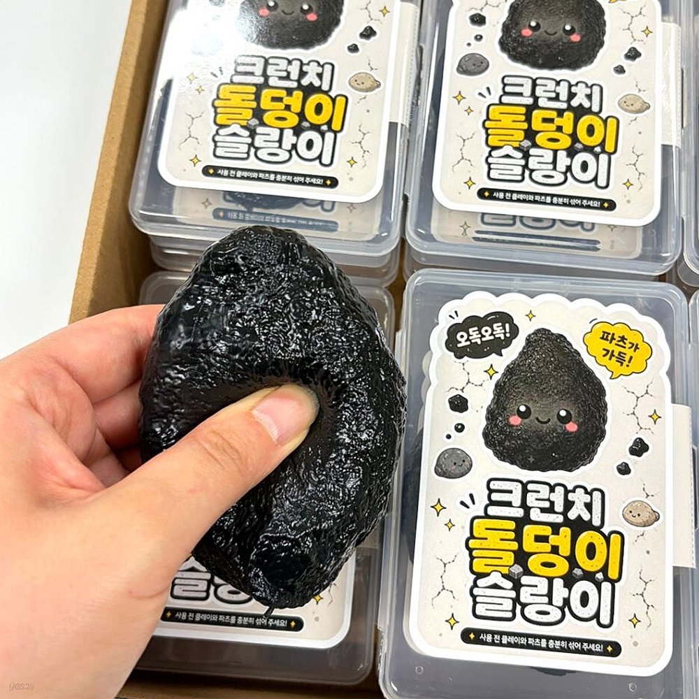 Hand holding and squeezing a black Charcoal Stone Crunch Squishy Slime Toy over clear plastic storage cases with cute Korean stone character labels, ideal stress relief sensory fidget toy and holiday stocking stuffer