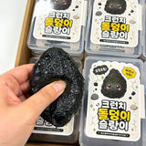 Hand holding and squeezing a black Charcoal Stone Crunch Squishy Slime Toy over clear plastic storage cases with cute Korean stone character labels, ideal stress relief sensory fidget toy and holiday stocking stuffer