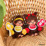 Two Monchhichi dolls with colorful accessories on a woven background